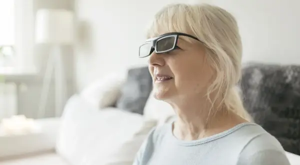 Une femme âgée porte des lunettes spéciales, peut-être dans une maison de retraite spécialisée pour les malvoyants, profitant d'un moment de confort.