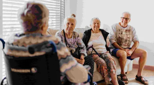 Quatre personnes âgées discutent sur un canapé. L'une est en fauteuil roulant. Le tarif du foyer logement senior est abordé.