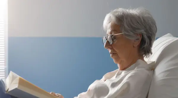 Une femme âgée, en **had et apa**, lit un livre confortablement installée dans un lit.