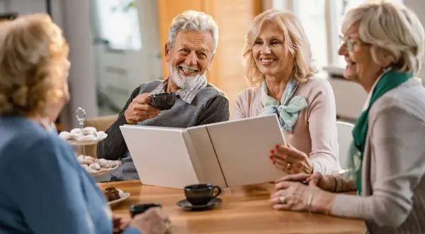 Quatre seniors souriants partagent un moment convivial autour d'un album. L'espérance de vie après entrée en EHPAD peut être belle et joyeuse.