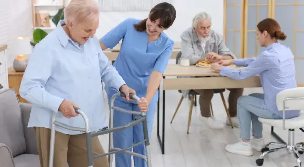 Une aide-soignante accompagne une résidente d'un EHPAD Alzheimer avec un déambulateur. Au fond, d'autres résidents sont à table.