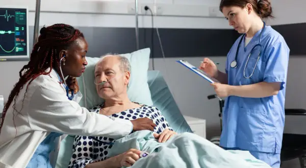 Soins attentifs pour une **hospitalisation à domicile cancer phase terminale**. Une infirmière écoute le cœur d'un patient âgé, une autre note des informations.
