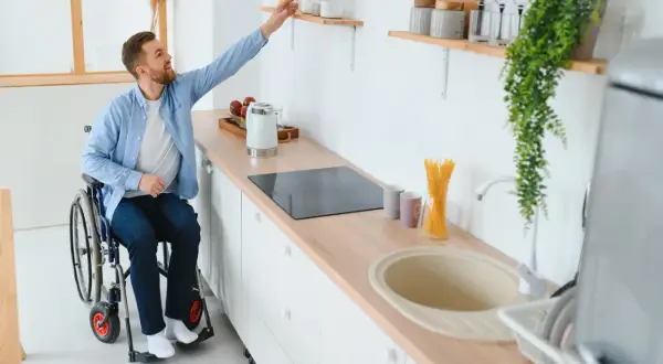 Un homme en fauteuil roulant attrape un objet sur une étagère dans une cuisine adaptée. Un exemple de priorité handicap logement social.