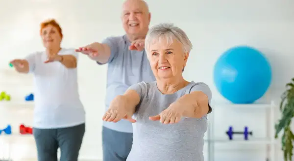 Trois seniors souriants participent à un exercice de gymnastique, bras tendus, dans une salle lumineuse.