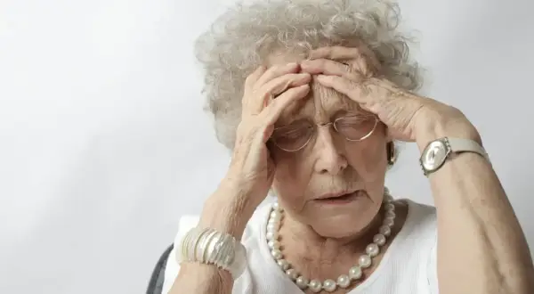 Une femme âgée, aux cheveux gris bouclés, se tient la tête, le regard perdu. Elle porte des lunettes, un collier de perles et un bracelet. Son expression suggère une détresse, peut-être liée à un stade avancé de la maladie d'Alzheimer.