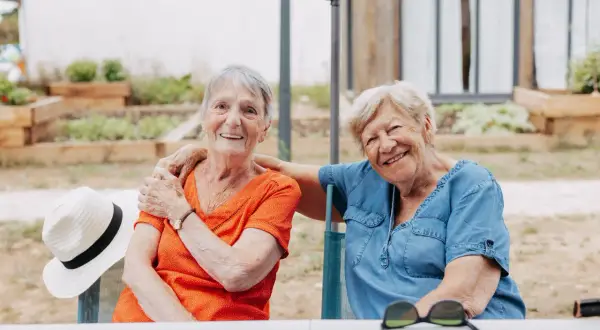 Deux femmes âgées souriantes, profitant de leur logement pour seniors en plein air. Une chapeau de paille est posé sur une chaise.