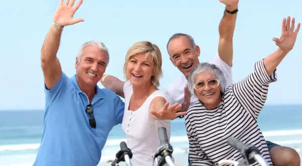 Quatre seniors joyeux agitent les mains, profitant d'une belle journée au bord de la mer, peut-être après une balade à vélo dans ce village senior bord de mer.