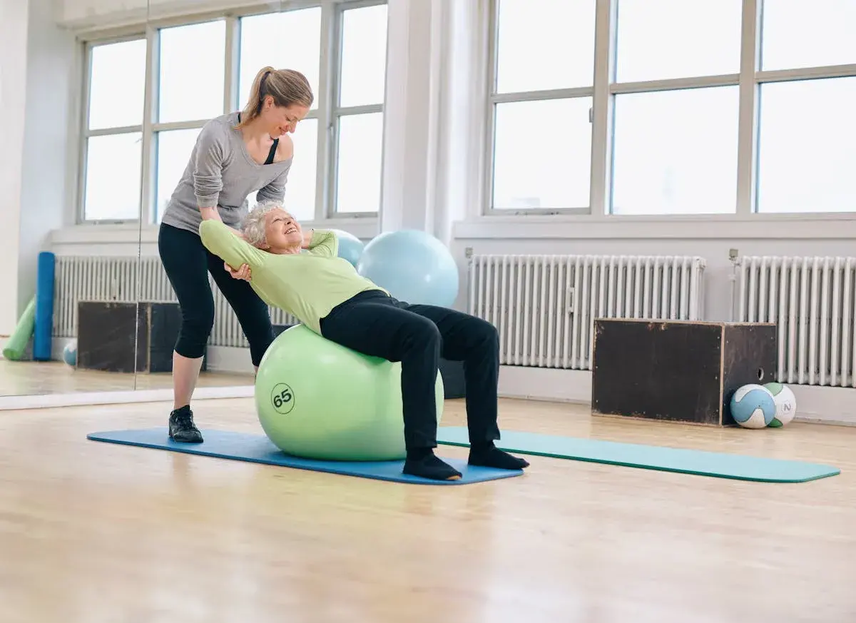 Une jeune femme aide une senior à faire des exercices sur un ballon de gym. Le **gym dos senior** est une activité bénéfique.