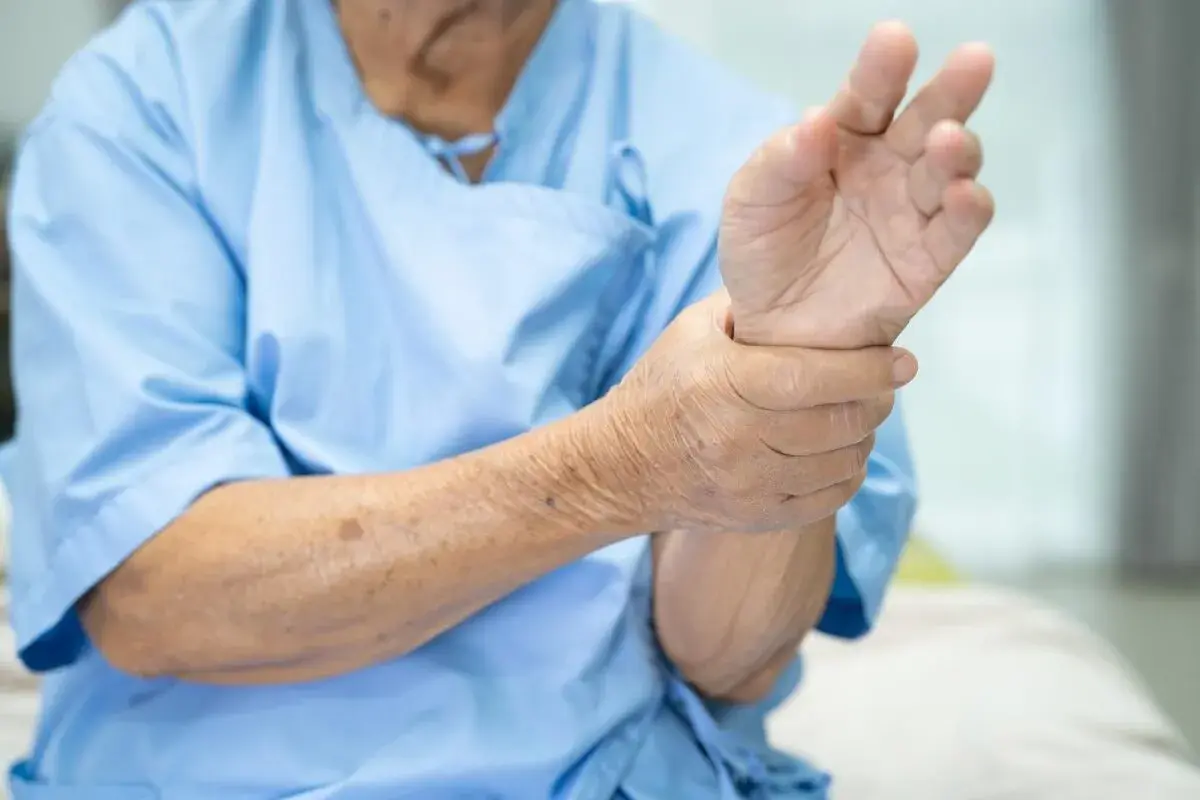 Main d'une personne âgée en blouse bleue, tenant son poignet, peut-être un pouce qui bouge tout seul.