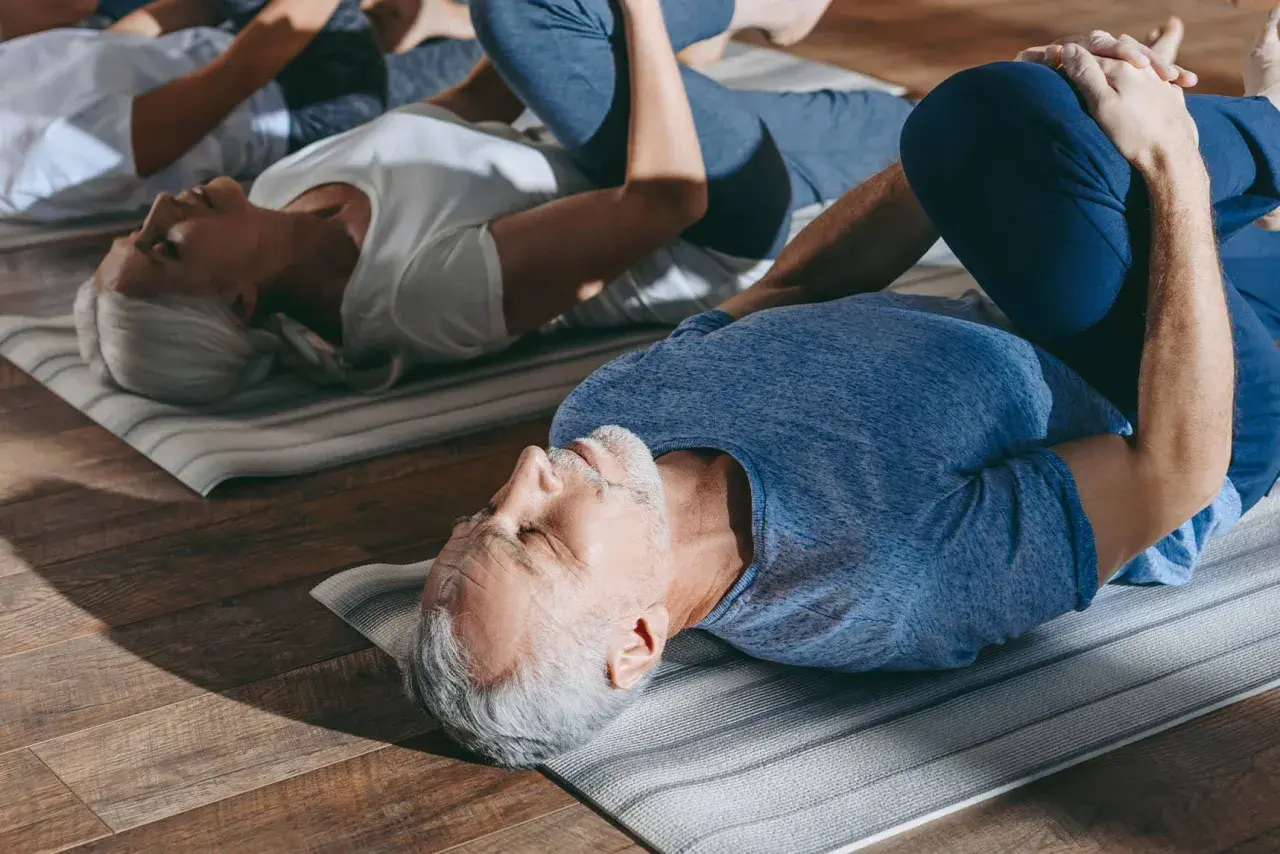 Des seniors pratiquent la gym douce sur des tapis. Un homme et une femme, les genoux pliés, se détendent.