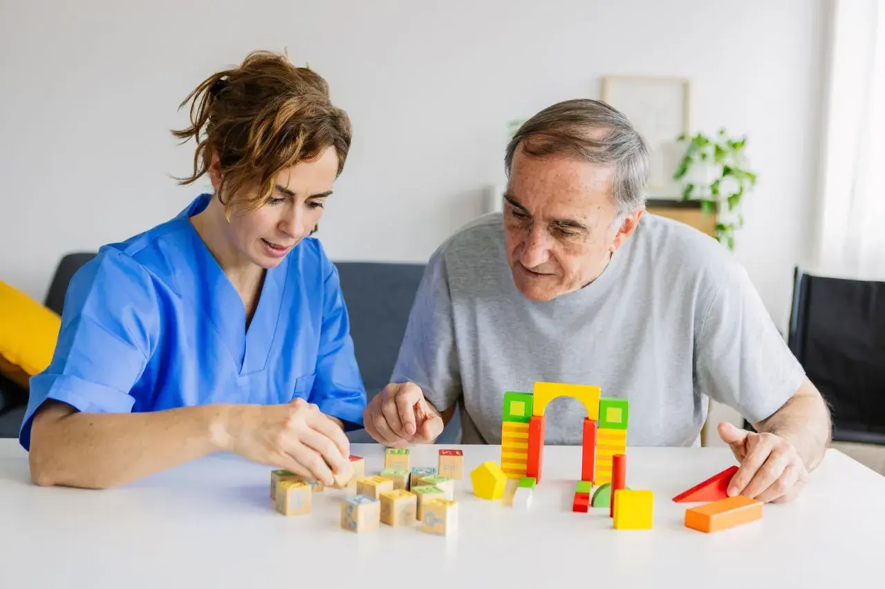 Une aide-soignante en blouse bleue joue avec des cubes en bois avec un homme âgé. Une scène de la vie quotidienne dans un établissement de soins, pas un pasa ehpad.