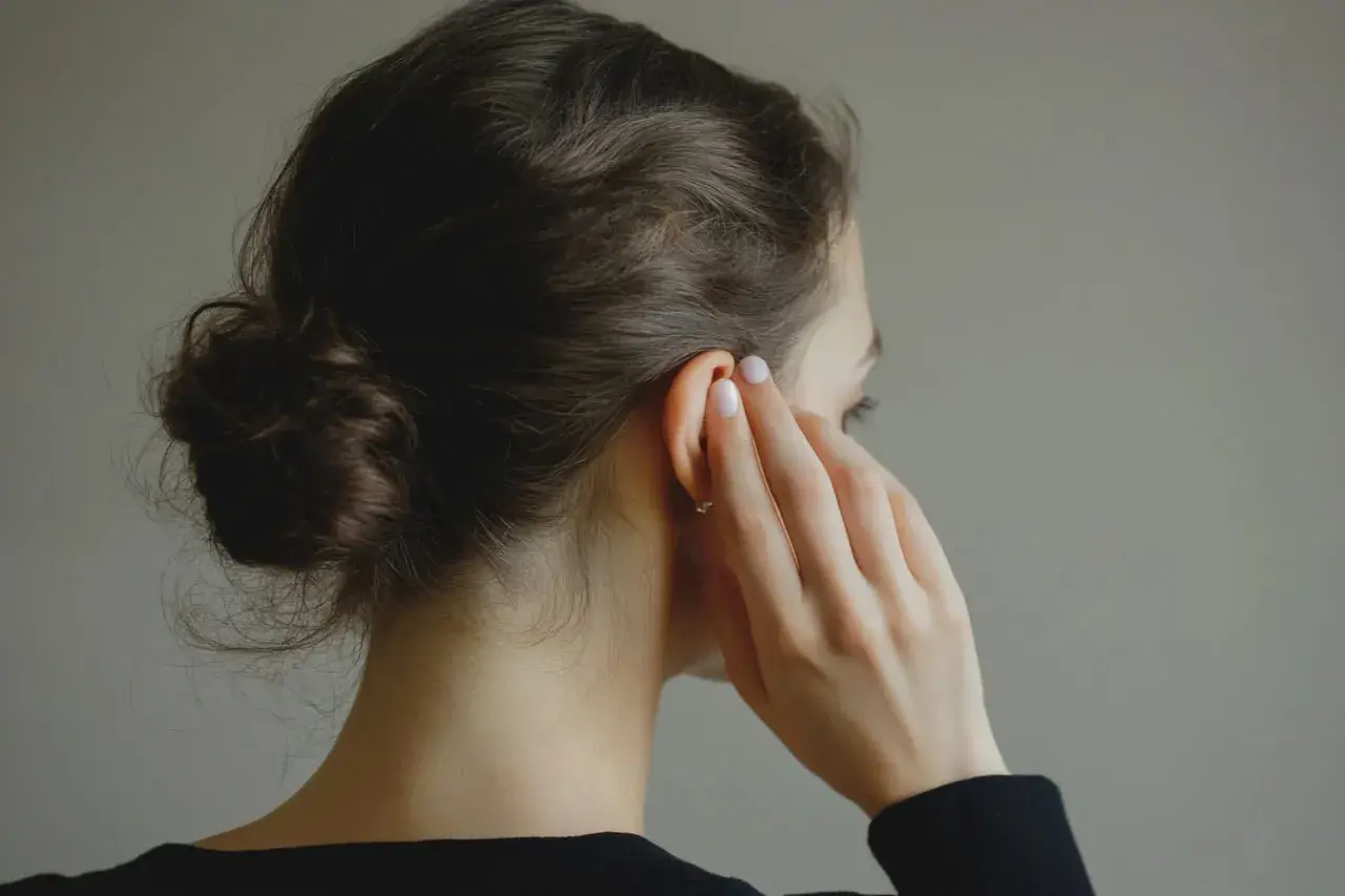 Une femme aux cheveux bruns en chignon touche son oreille, suggérant une solution naturelle comme l'huile d'amande pour les bouchons d'oreille.