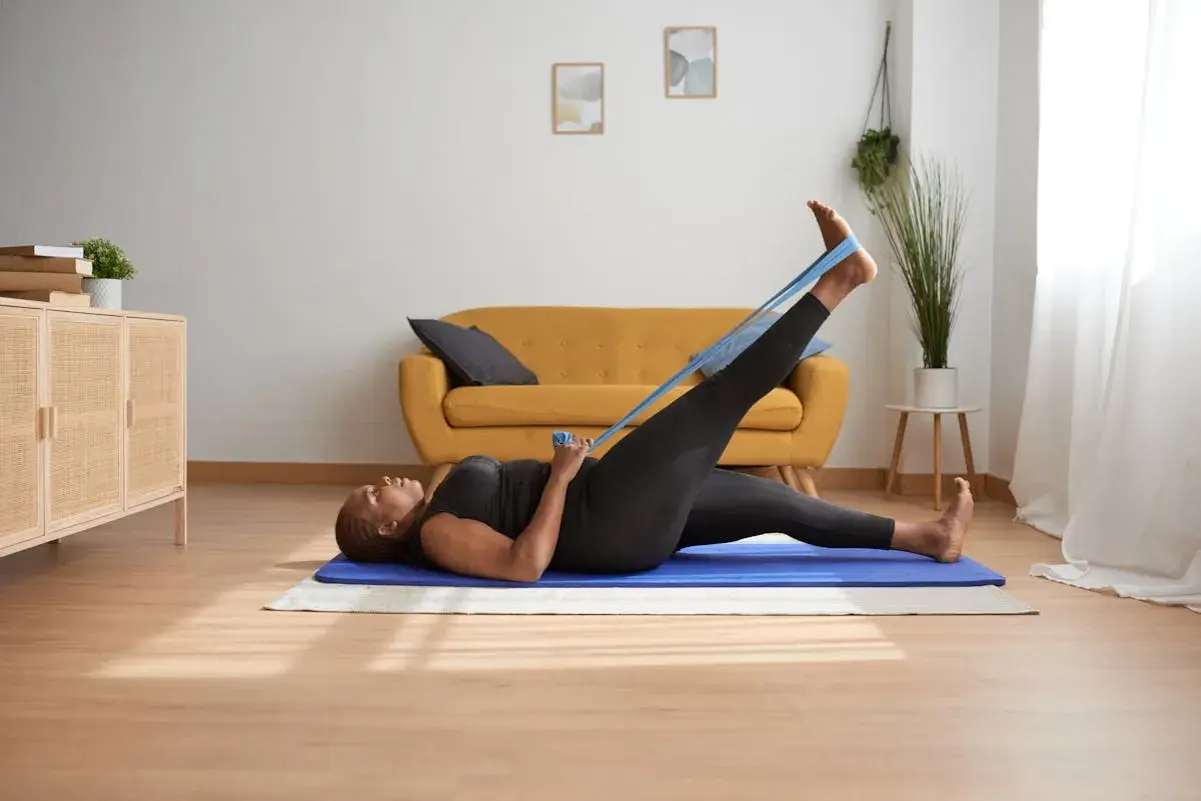 Femme faisant des exercices de gymnastique d'entretien avec une bande élastique, allongée sur un tapis bleu dans un salon lumineux.