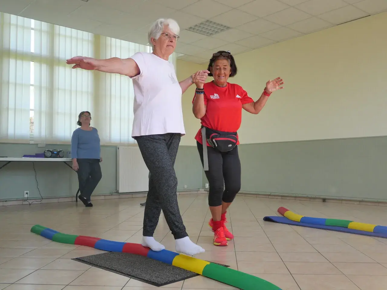 Deux seniors s'exercent sur un parcours équilibre, bras écartés pour maintenir leur stabilité. Une troisième femme observe.