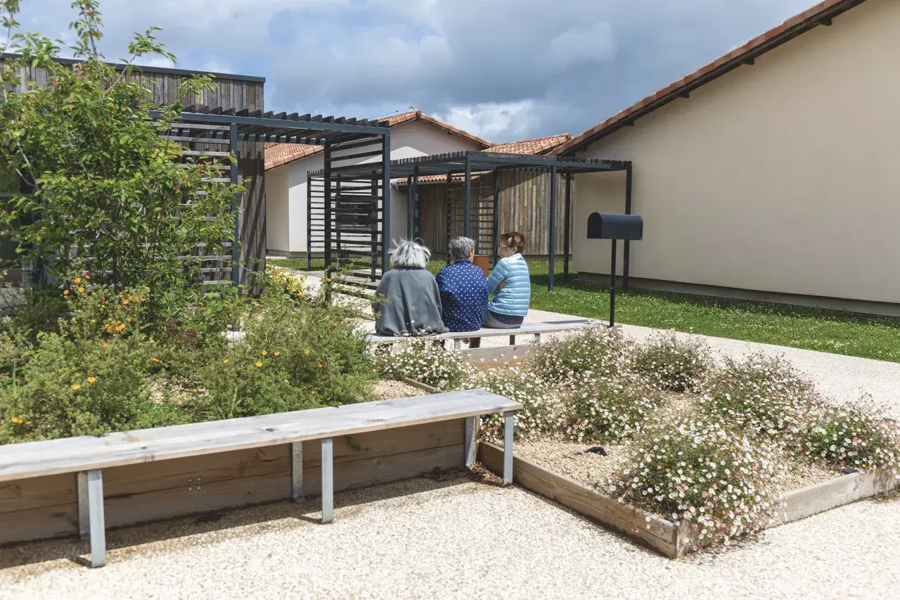 Trois femmes assises sur un banc dans un jardin fleuri, devant des maisons modernes. Un lieu de vie paisible pour un village Alzheimer à Dax.