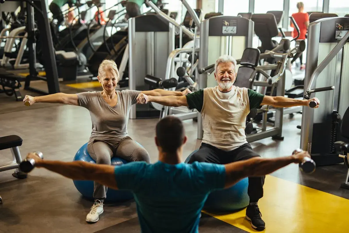Un couple de retraités souriants fait un exercice swiss ball avec des haltères, guidé par un coach.