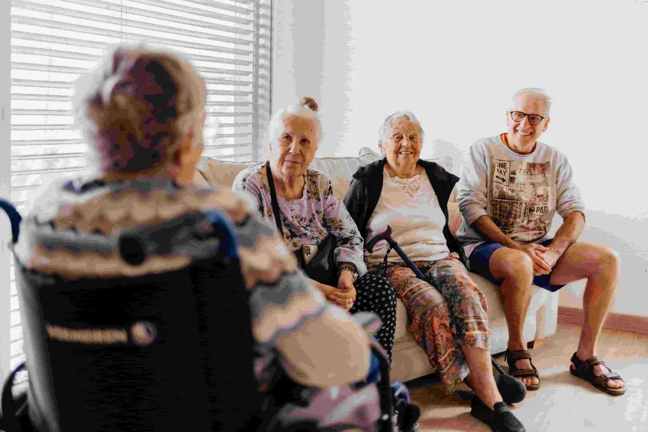 Quatre personnes âgées discutent sur un canapé. L'une est en fauteuil roulant. Le tarif du foyer logement senior est abordé.