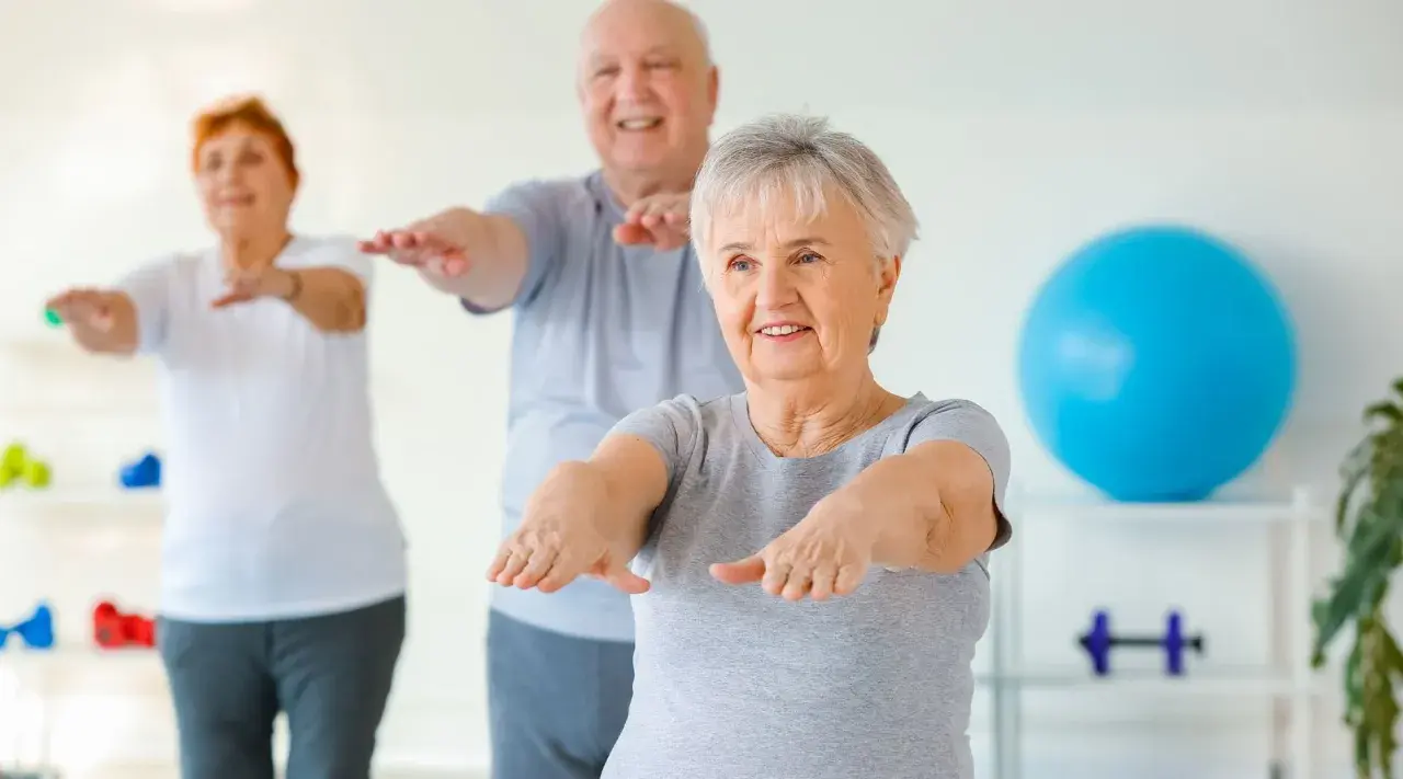 Trois seniors souriants participent à un exercice de gymnastique, bras tendus, dans une salle lumineuse.
