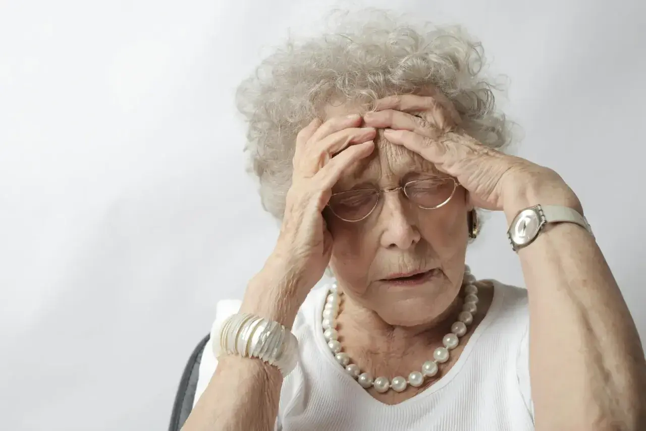 Une femme âgée, aux cheveux gris bouclés, se tient la tête, le regard perdu. Elle porte des lunettes, un collier de perles et un bracelet. Son expression suggère une détresse, peut-être liée à un stade avancé de la maladie d'Alzheimer.