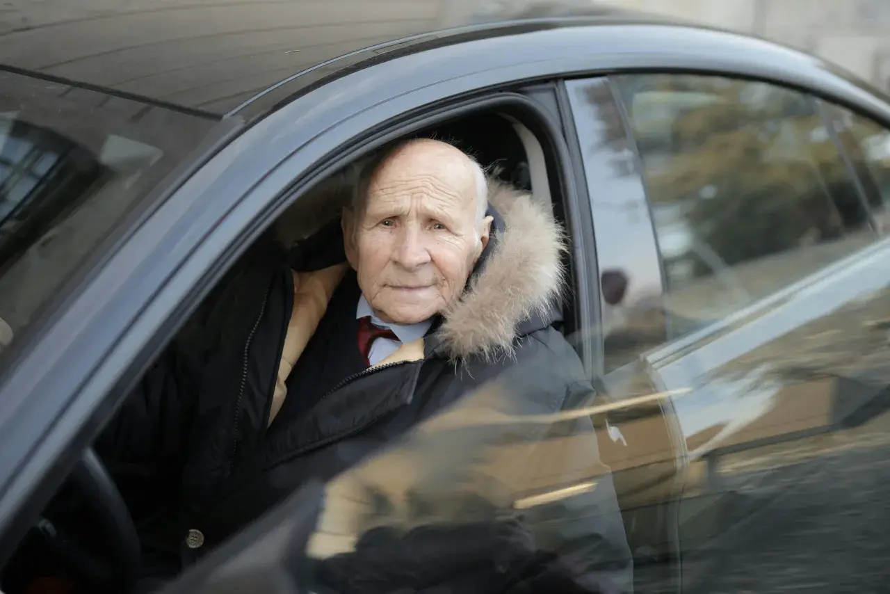 Un homme âgé, un senior à partir de quel âge, porte une veste d'hiver avec un col en fourrure, assis dans une voiture.