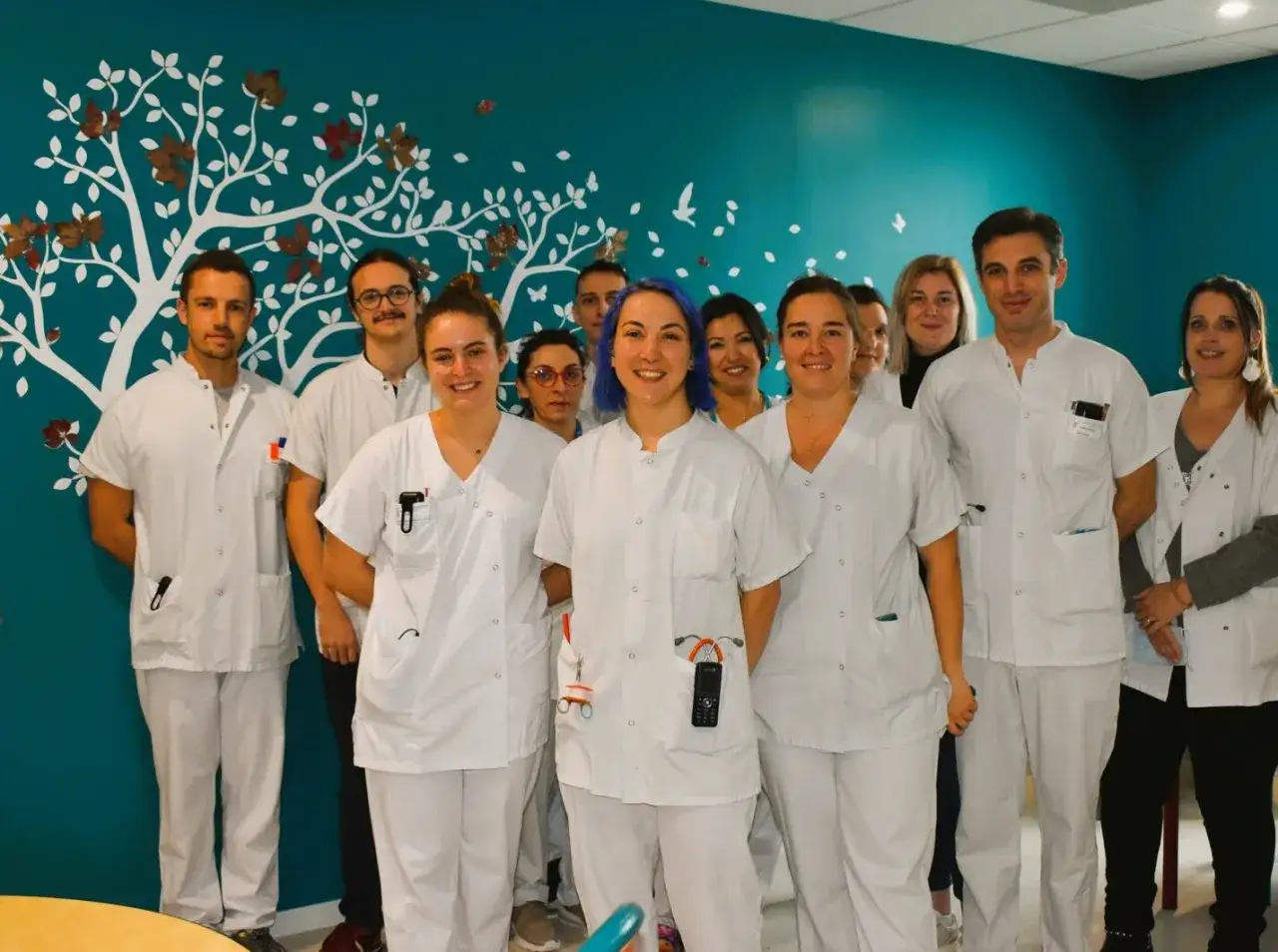 Une &eacute;quipe souriante de professionnels de sant&eacute; en blouse blanche pose devant un mur d&eacute;cor&eacute; d'un arbre stylis&eacute;, au sein de l'usld hopital.