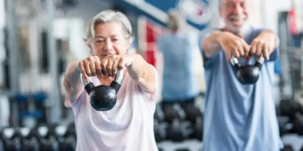 Un couple de retrait&eacute;s souriants participe activement &agrave; un programme de mise en forme 60 ans et plus, soulevant des kettlebells avec d&eacute;termination.