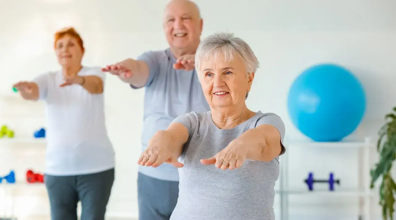 Trois seniors souriants font un exercice assis sur une chaise, bras tendus.