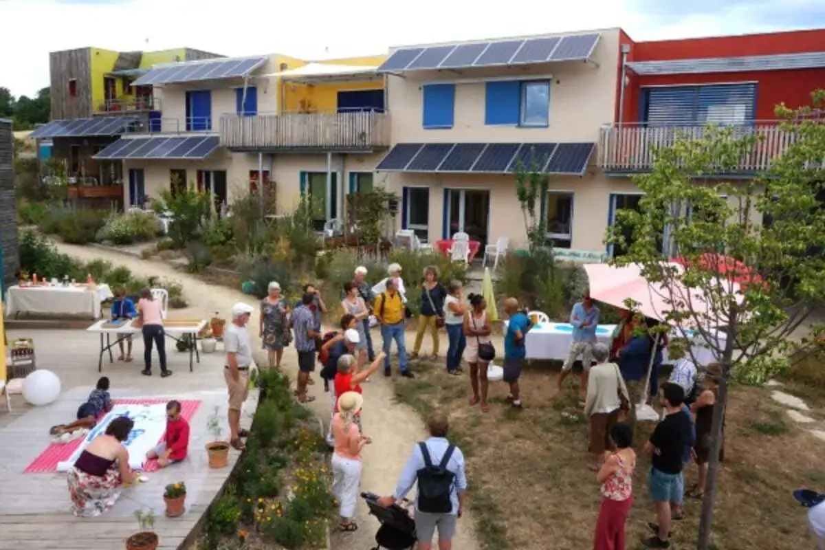 Un groupe de personnes profite d'un rassemblement ensoleillé devant des maisons modernes équipées de panneaux solaires, illustrant un projet d'habitat participatif seniors.