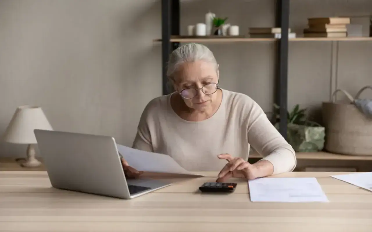 Une femme &acirc;g&eacute;e, lunettes sur le nez, calcule des chiffres sur une calculatrice, un ordinateur portable ouvert &agrave; c&ocirc;t&eacute;. Elle semble g&eacute;rer des documents importants, peut-&ecirc;tre pour son apa en ehpad.