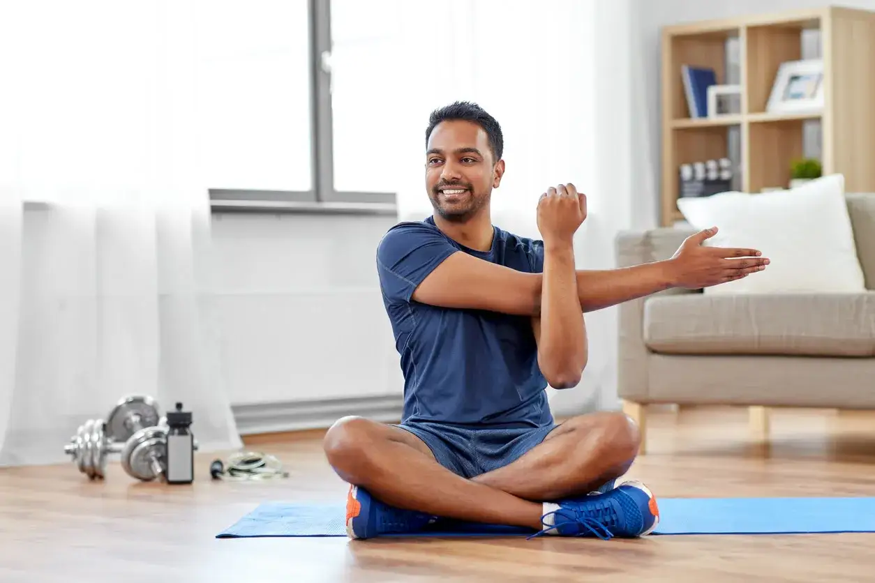 Un homme s'étire sur un tapis bleu, prêt pour un gym facile pour débutant. Des haltères et une corde à sauter sont à proximité.