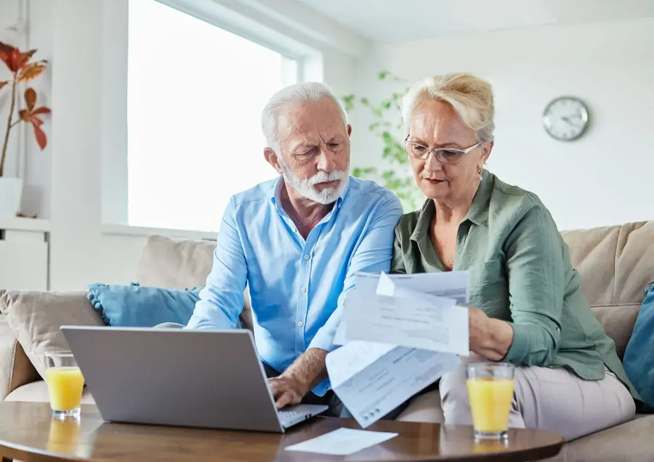 Couple &acirc;g&eacute; examinant des documents, peut-&ecirc;tre pour une exon&eacute;ration taxe fonci&egrave;re 2026. Ils sont sur un canap&eacute; avec un ordinateur portable et des boissons.