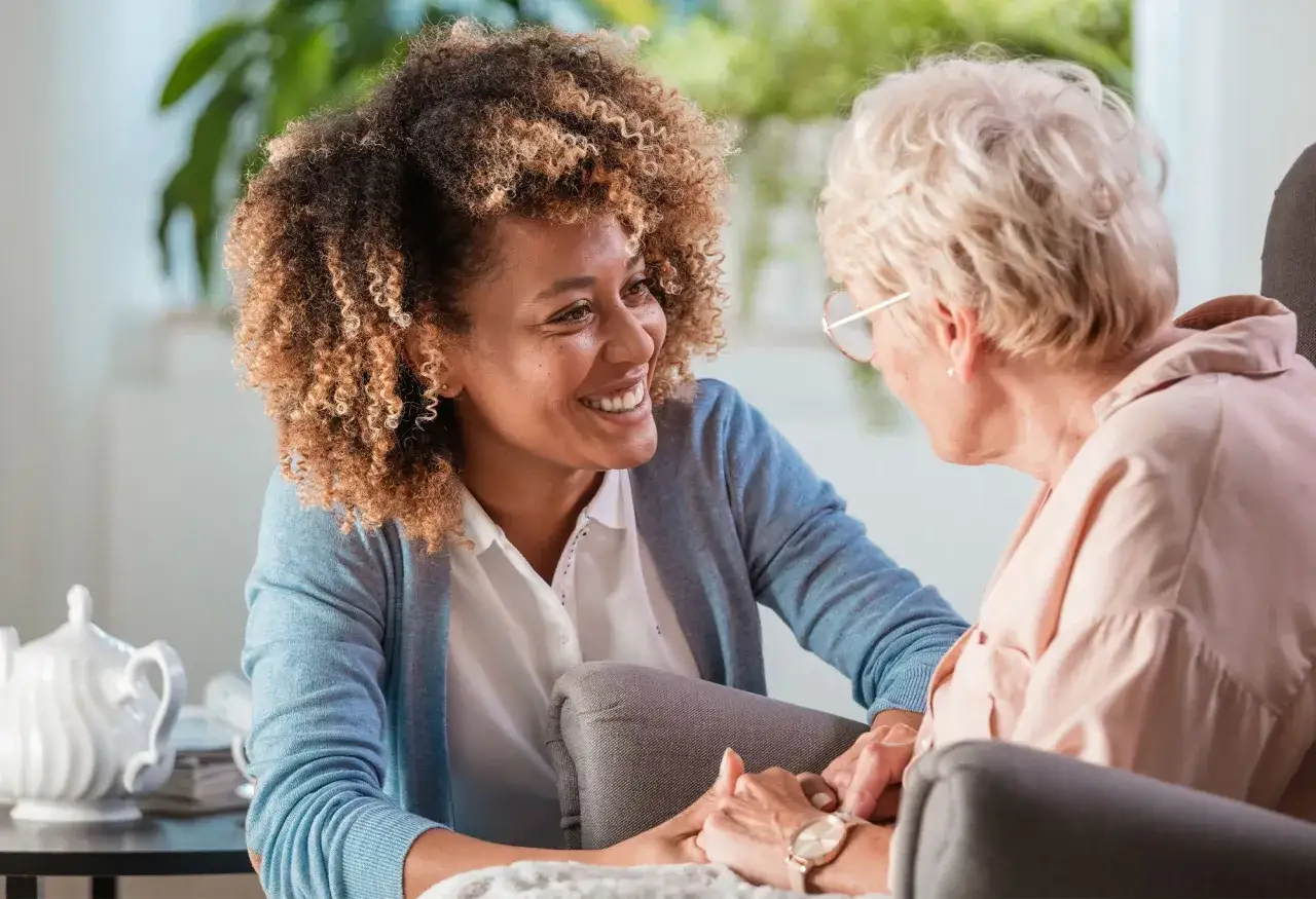 Une aide souriante discute avec une personne &acirc;g&eacute;e, main dans la main. Un moment de complicit&eacute; qui illustre un plan d'aide personnalis&eacute; pour personne &acirc;g&eacute;e.
