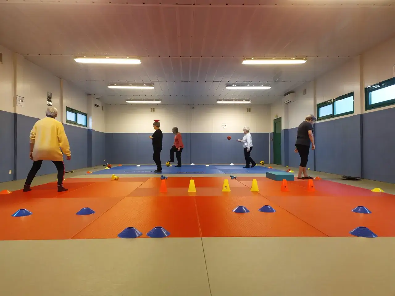 Des seniors s'entraînent sur un parcours d'équilibre avec des cônes et des ballons dans un gymnase.