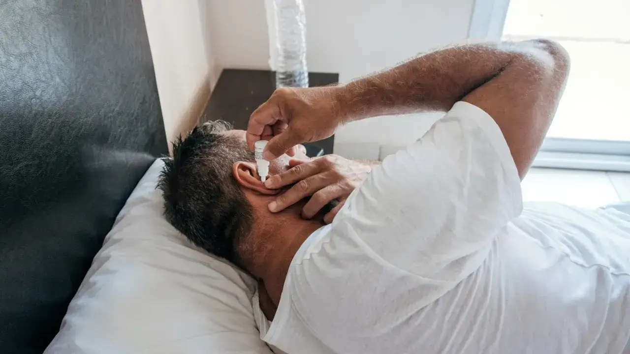 Homme allong&eacute; sur un oreiller, appliquant de l'huile d'amande dans son oreille pour soulager des bouchons.