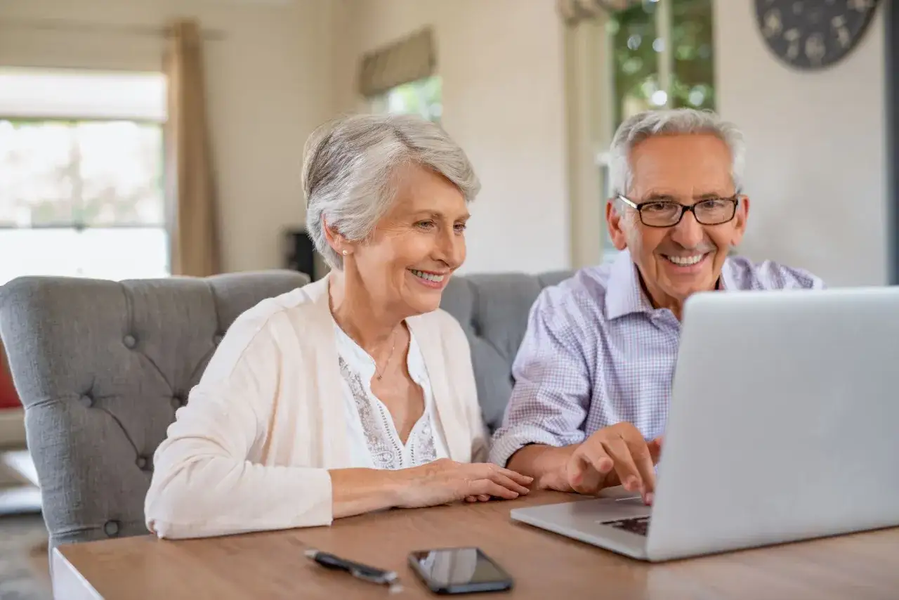 Couple âgé souriant devant un ordinateur portable, peut-être en train de consulter le tarif foyer logement senior.