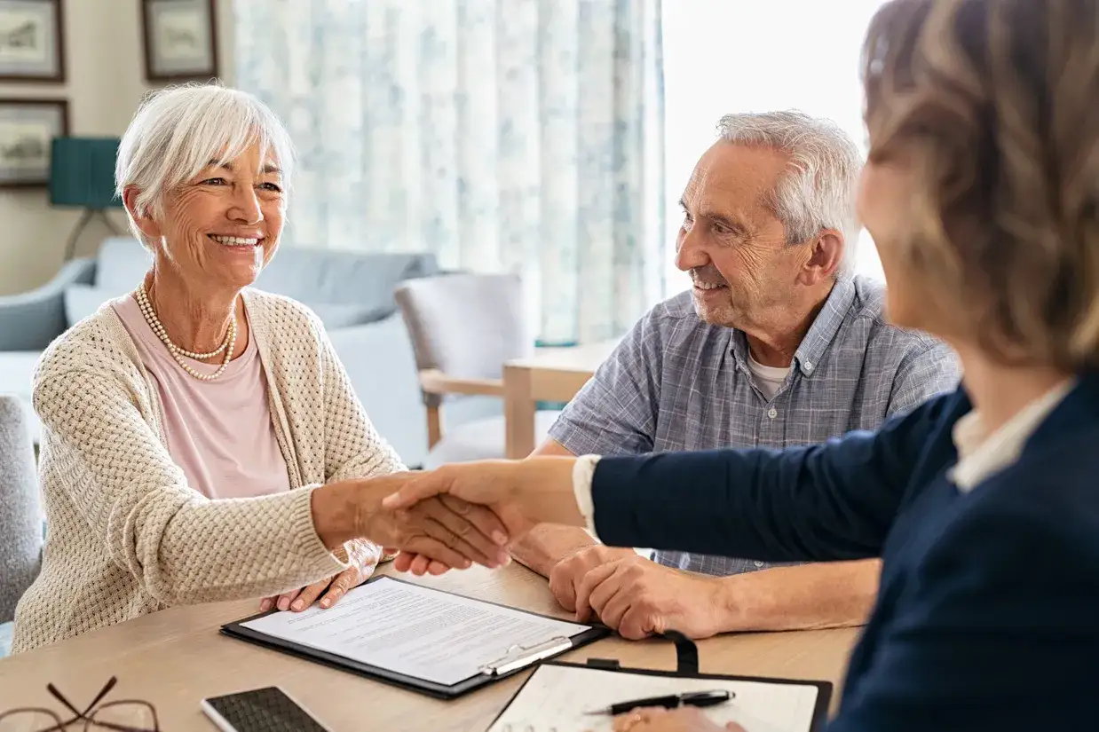 Un couple &acirc;g&eacute; serre la main d'une conseill&egrave;re, souriant. Ils discutent peut-&ecirc;tre d'une maison de retraite gratuite.