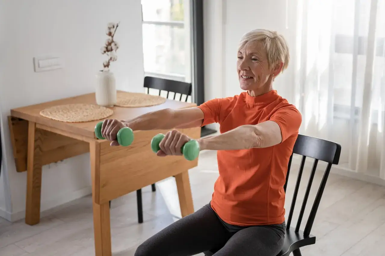 Femme souriante faisant de la gym douce avec des haltères verts, assise sur une chaise noire.