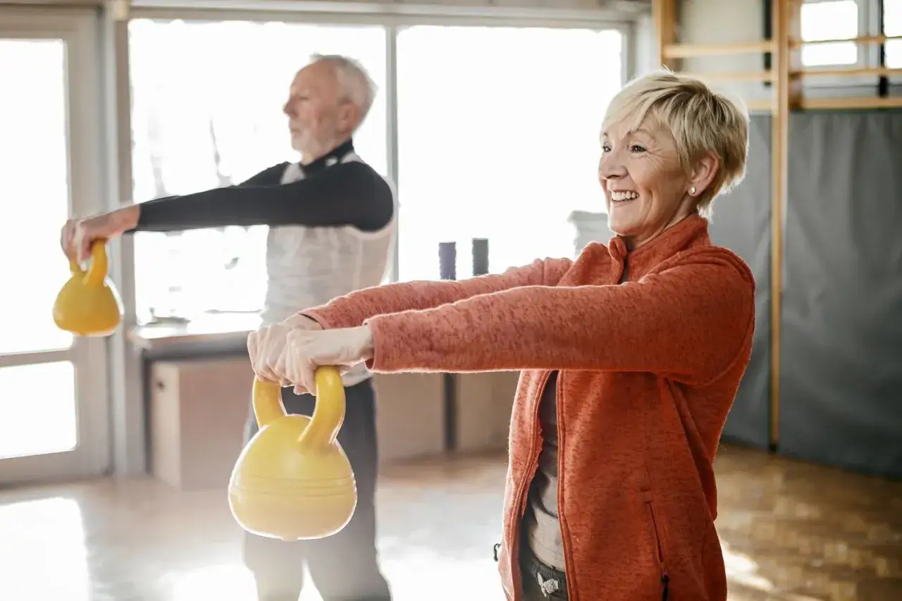 Un couple souriant fait des charges pour musculation à 70 ans, soulevant des kettlebells jaunes dans une salle de sport lumineuse.
