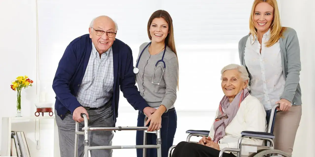 Un homme âgé utilise un déambulateur, une femme en fauteuil roulant est assistée par deux soignantes. Une scène de soutien et de soins, loin de l'hospitalisation.