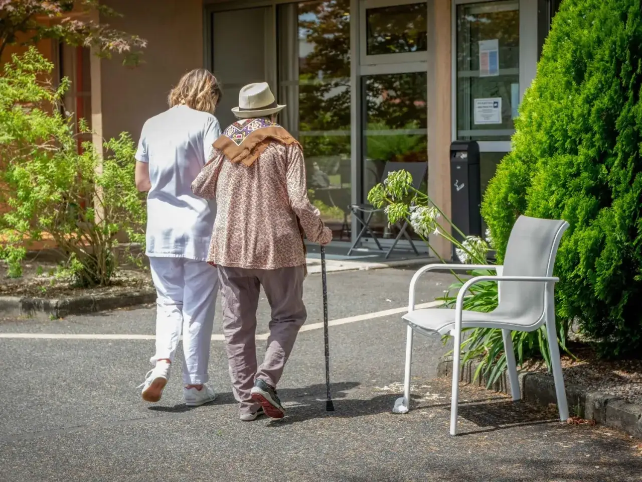 Une aide-soignante accompagne une personne âgée, en chapeau et canne, dans le cadre d'une curatelle aménagée.