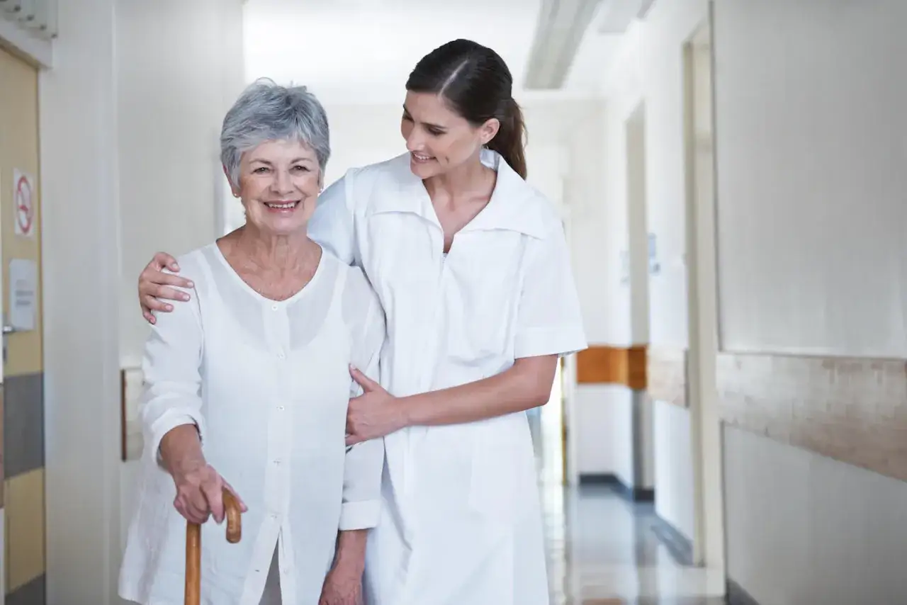 Une aide-soignante soutient une r&eacute;sidente souriante dans un couloir d'EHPAD. L'image &eacute;voque l'esp&eacute;rance de vie apr&egrave;s entr&eacute;e en ehpad, gr&acirc;ce &agrave; des soins attentifs.
