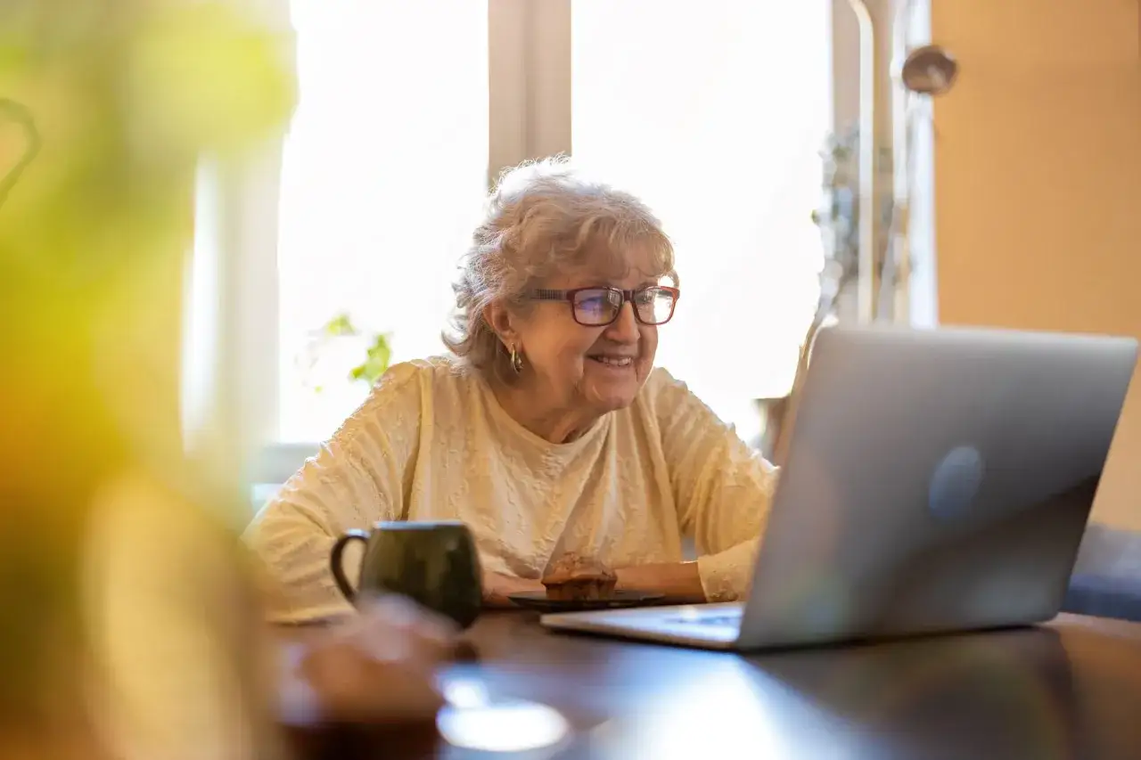 Une dame âgée souriante utilise son ordinateur portable, peut-être pour une aide à domicile ou un suivi GIR 2. Une tasse et un muffin sont à proximité.