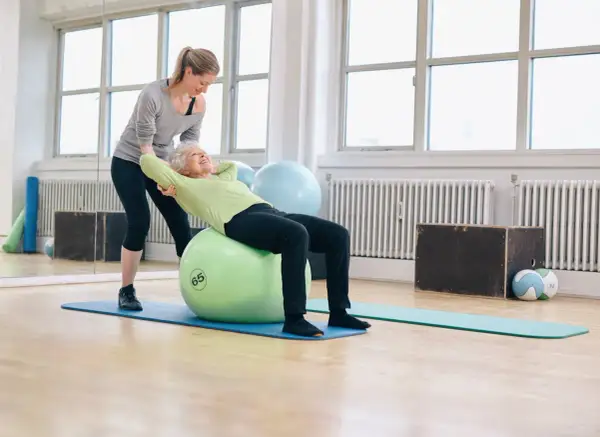 Une jeune femme aide une senior à faire des exercices sur un ballon de gym. Le **gym dos senior** est une activité bénéfique.