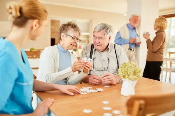 Des aînés assemblent un puzzle dans une maison de retraite gratuite, aidés par une aide soignante.