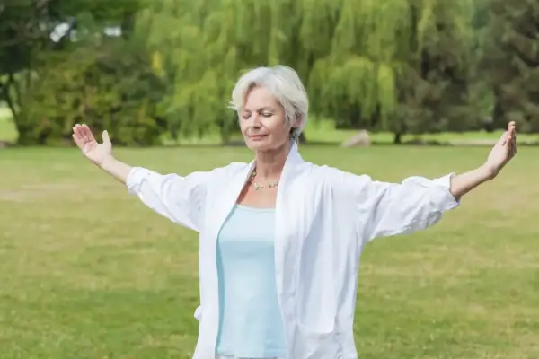 Femme d'âge mûr pratiquant le mouvement qi gong dans un parc, bras écartés, yeux fermés, en harmonie avec la nature.