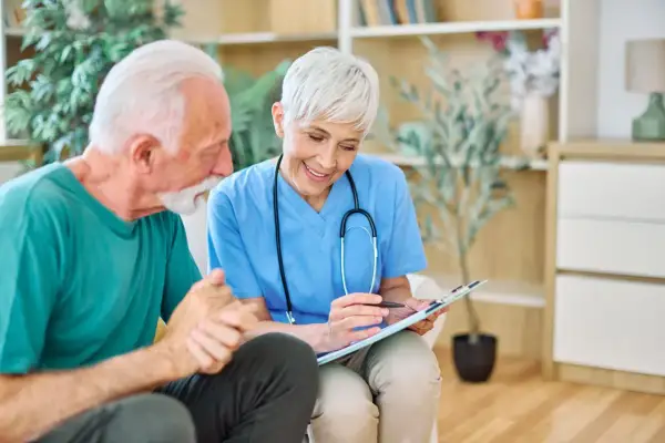 Une aide-soignante discute avec un patient âgé, lui expliquant son plan de soins. Elle tient un dossier, et le patient semble attentif. La question "combien d'heures d'aide pour un gir 3" est au cœur de leur échange.