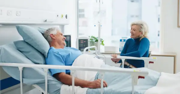Un homme âgé, en chemise d'hôpital, sourit à une femme assise à son chevet. Une scène réconfortante d'apa et d'hospitalisation.