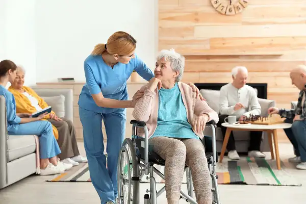 Une aide-soignante soutient une femme en fauteuil roulant, tandis que d'autres résidents jouent aux échecs. L'image évoque le soin et la transmission, comme une succession de moments de vie.