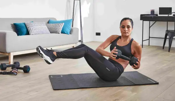 Femme faisant des torsions russes avec haltère, un exercice de gym facile pour débutant, dans un salon moderne.