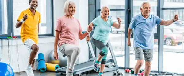 Quatre seniors souriants participent à un cours de gym, faisant des exercices dynamiques dans une salle lumineuse.
